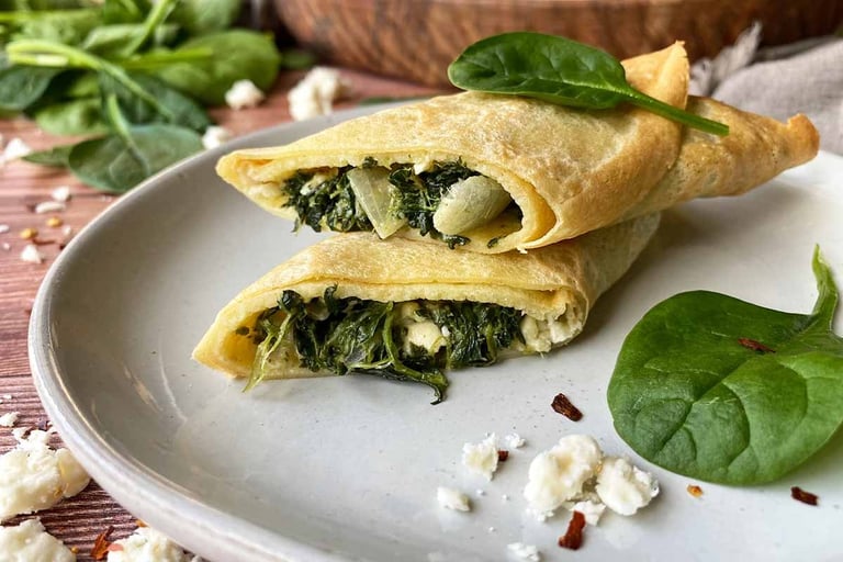 Two crepes filled with spinach and cheese on a white plate, garnished with fresh spinach leaves and crumbled cheese